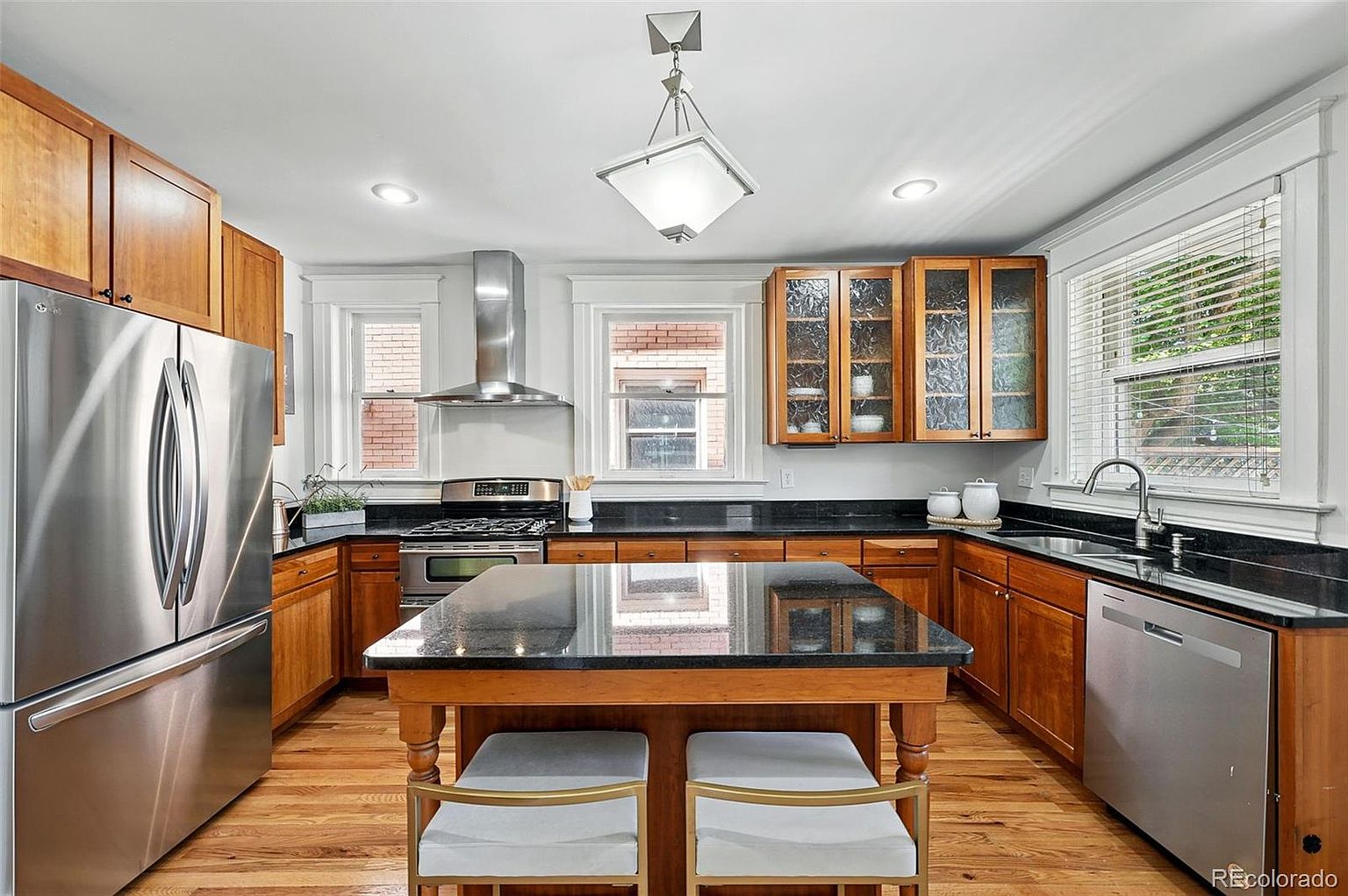 This is a well-lit kitchen featuring wooden cabinetry, stainless steel appliances, and a dark countertop. A kitchen island with seating is centrally located, and natural light streams in through the windows. The overall impression is a blend of traditional and modern elements.