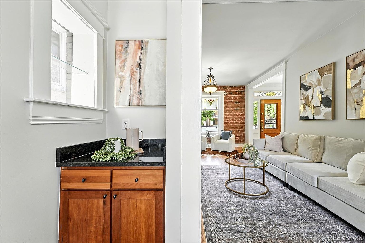 This interior shot showcases a stylish living room with a neutral color palette. A large sectional sofa anchors the space, complemented by a patterned area rug and a round coffee table. Artwork adorns the walls, and a glimpse into an adjacent room reveals exposed brick and a welcoming entryway, creating a warm and inviting atmosphere.