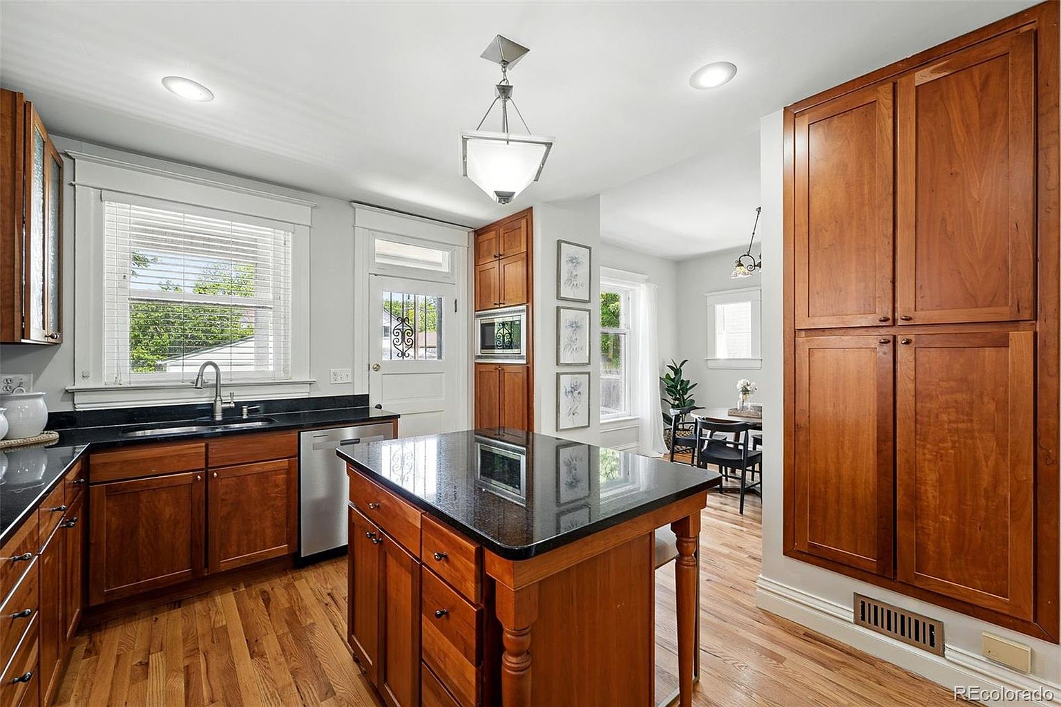 This is a well-lit kitchen featuring wooden cabinetry, black countertops, and hardwood floors. A central island with a black countertop provides additional workspace, and stainless steel appliances are visible. The kitchen has a warm and inviting feel, with natural light coming through the windows.