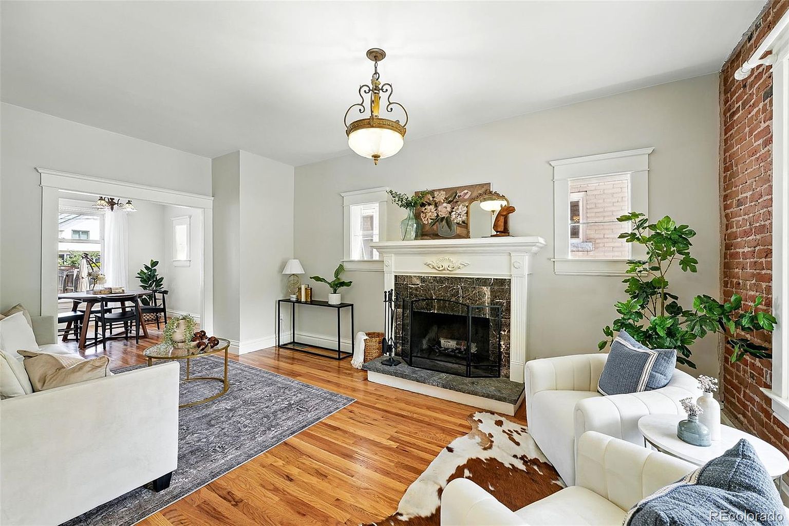 This is an interior shot of a living room featuring hardwood floors, a fireplace with a decorative mantel, and neutral-colored walls. The room is furnished with white armchairs, a patterned area rug, and a glass-topped coffee table. Natural light streams in through the windows, creating a bright and inviting atmosphere.