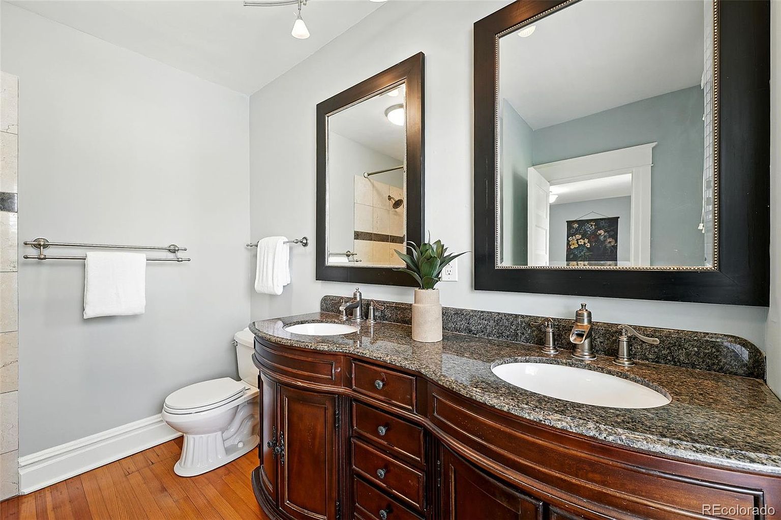 This is a primary bathroom featuring a double vanity with a dark wood finish and granite countertop. Two framed mirrors hang above the sinks, and the walls are painted a light blue. A toilet is visible to the left, and hardwood floors add warmth to the space.