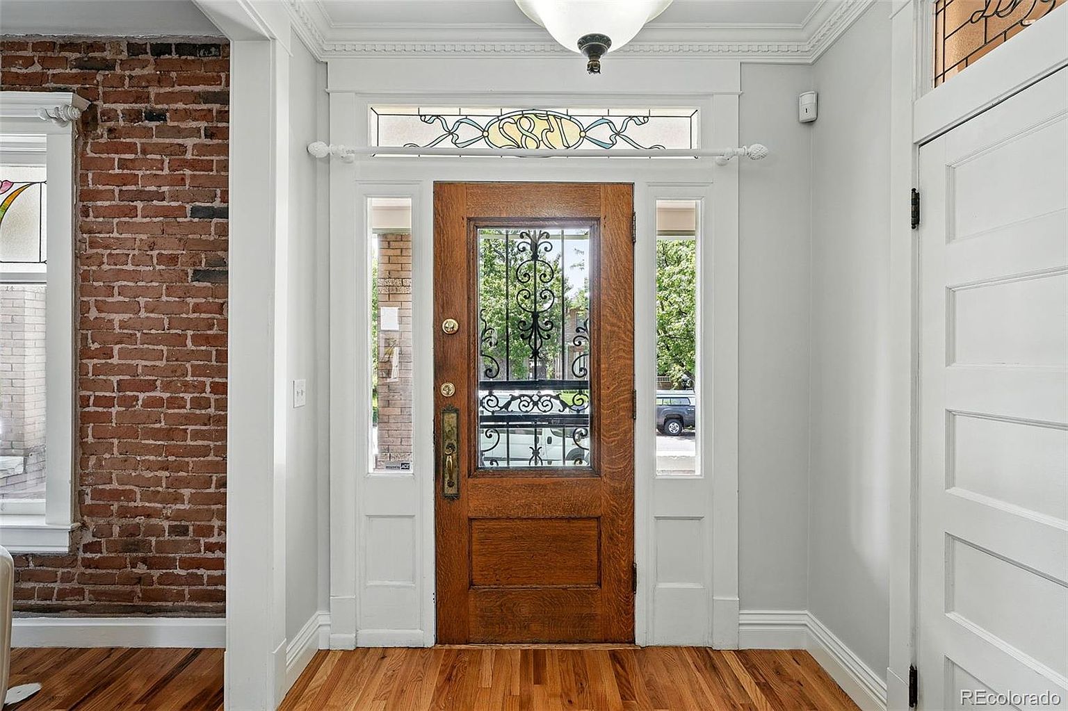 This interior shot showcases a hallway with a striking wooden front door featuring decorative ironwork and sidelights. The hallway is characterized by a blend of exposed brick and painted walls, complemented by hardwood flooring. A decorative transom window above the door adds an elegant touch, enhancing the overall charm of the entryway.