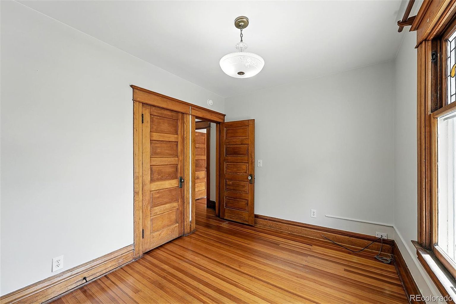 This is an interior shot of a bedroom featuring hardwood floors and light gray walls. The room has two wooden doors, one of which is a sliding door, and a vintage-style light fixture hangs from the ceiling. The room appears clean and well-maintained, offering a classic aesthetic.