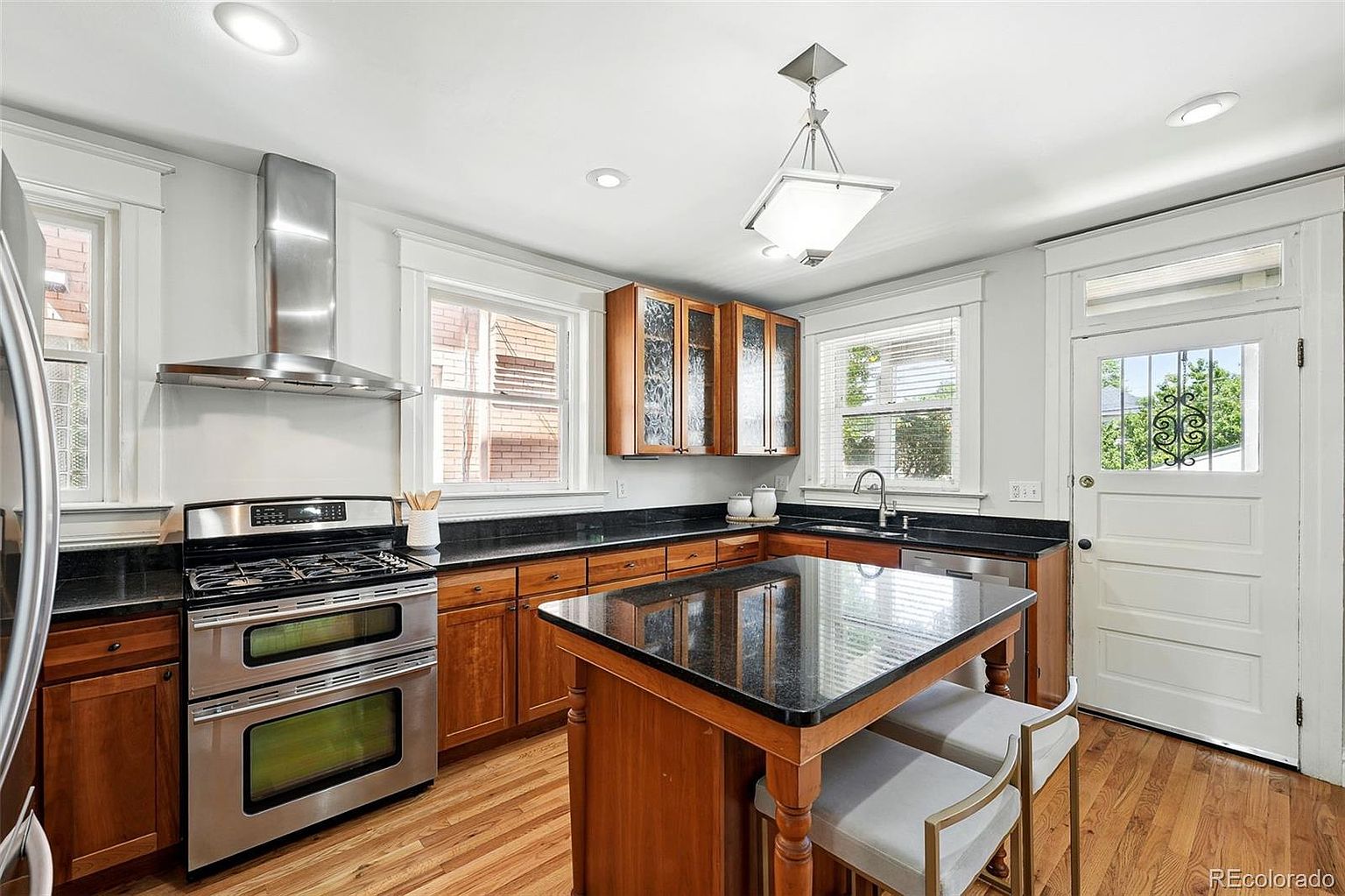 This is a well-lit kitchen featuring wooden cabinetry with black countertops and stainless steel appliances. A central island with a black countertop and bar stools provides additional workspace and seating. The hardwood flooring and natural light from the windows create a warm and inviting atmosphere.