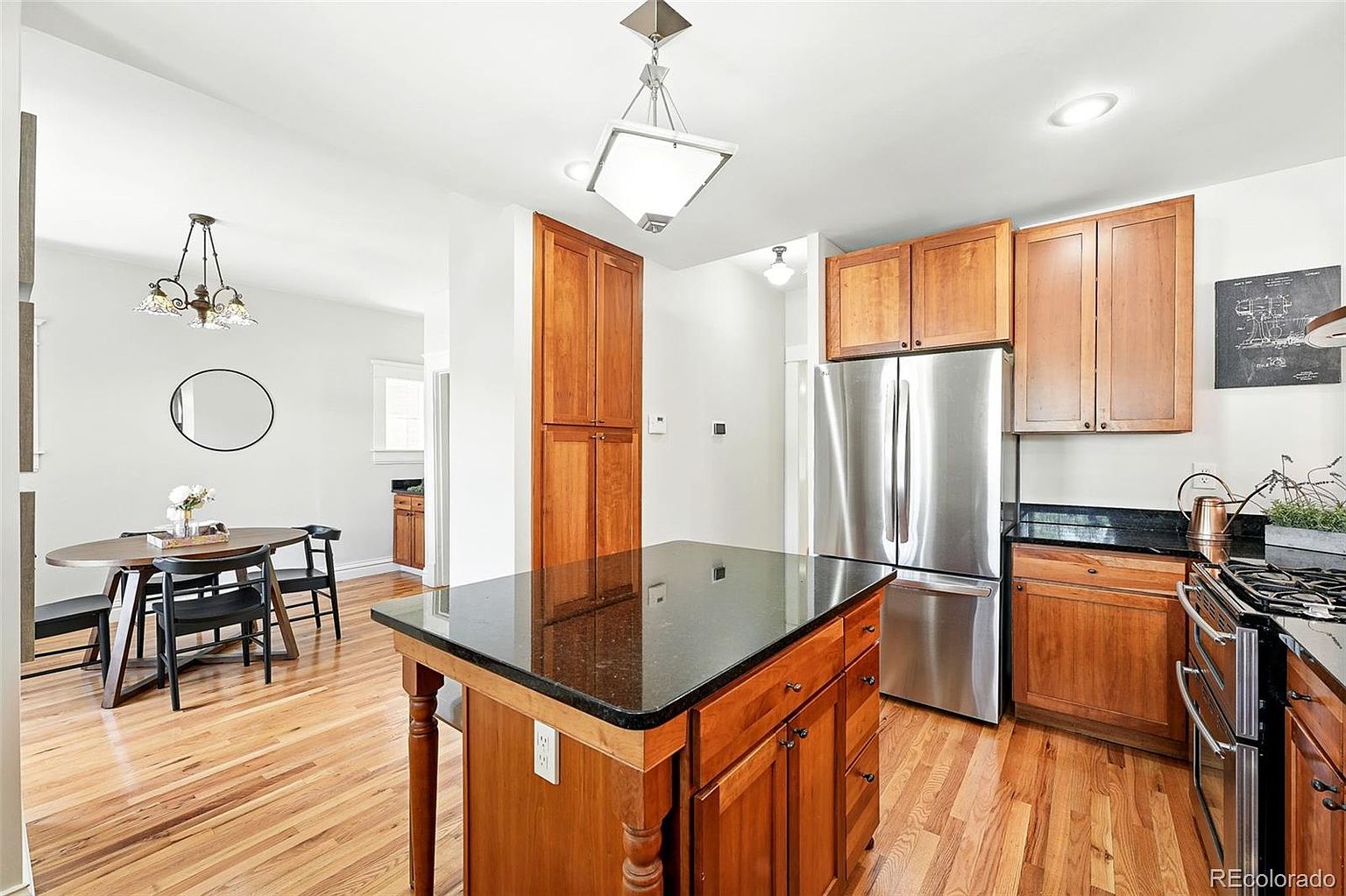 This is a well-lit kitchen featuring wooden cabinetry, stainless steel appliances, and a dark countertop island. The kitchen has hardwood floors and is connected to a dining area with a round table and black chairs. The overall impression is warm and inviting, with a blend of traditional and modern elements.