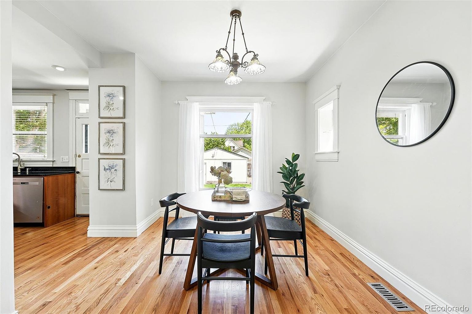 This is a bright and airy dining room featuring a round wooden table surrounded by four black chairs. Natural light floods the space through a window dressed with sheer white curtains, complemented by a decorative chandelier overhead. A round mirror hangs on the wall, adding depth to the room, while hardwood floors contribute to the warm and inviting atmosphere.