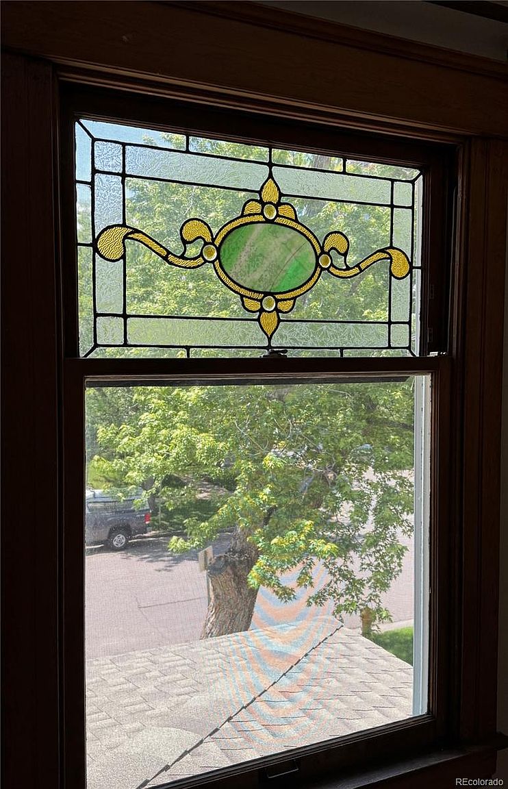 The image showcases a beautiful stained glass window with an ornate design, set within a dark wood frame. The window is divided into two sections, with the upper part featuring the stained glass and the lower part offering a view of the outside, including a tree and a glimpse of a roof. The overall impression is one of vintage charm and craftsmanship.