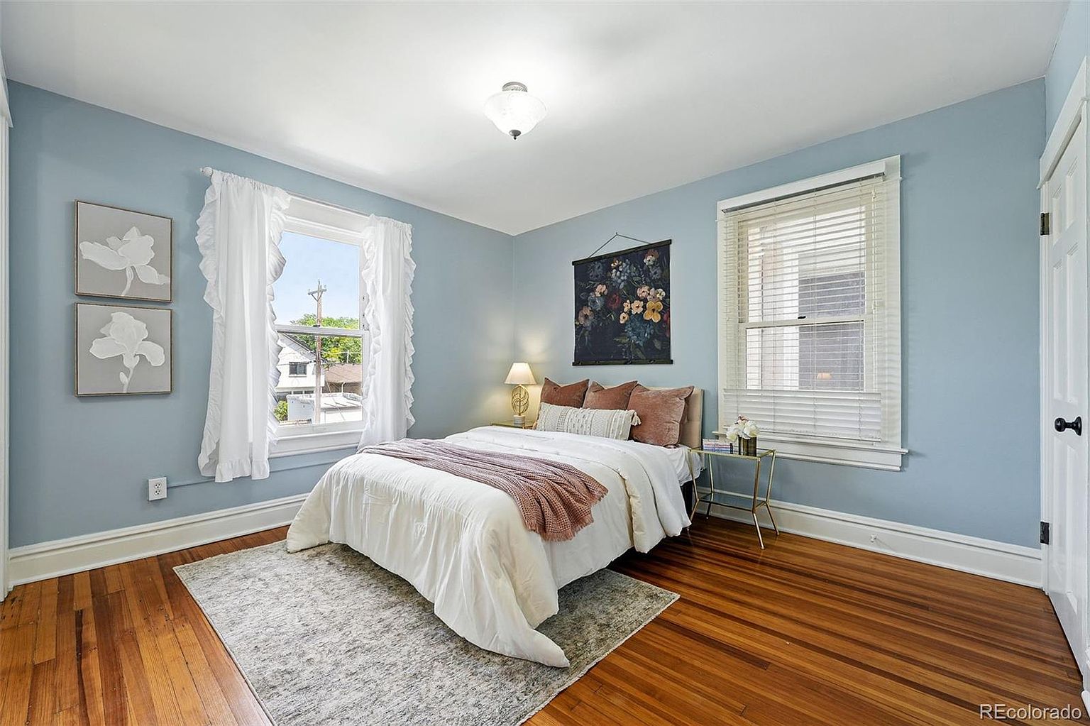This is a cozy bedroom featuring light blue walls, hardwood floors, and a neutral-toned rug. A bed with white linens and mauve accents is centered in the room, complemented by floral artwork and soft window treatments. The room has a bright and airy feel, enhanced by natural light from two windows.
