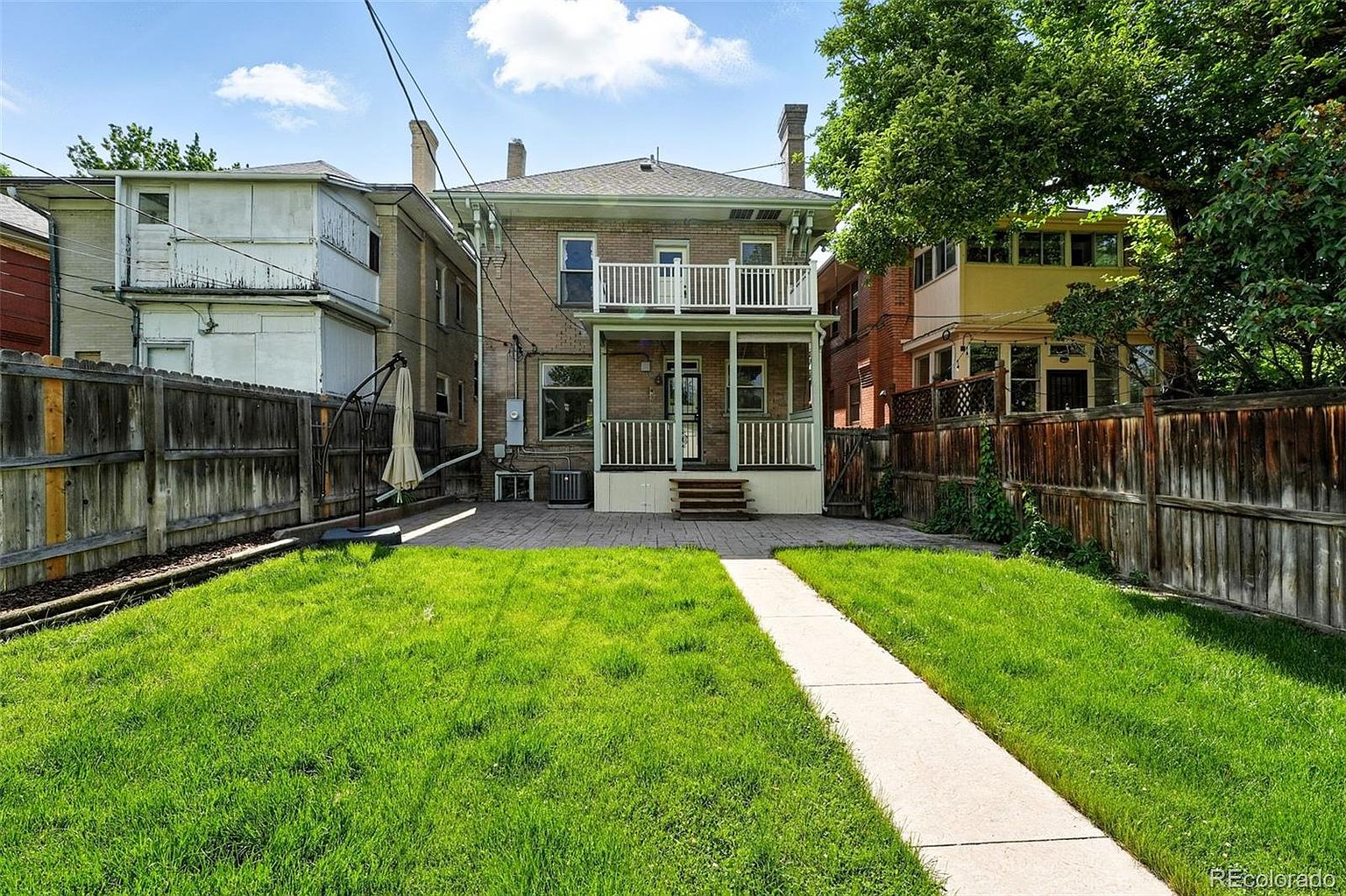 This is a rear view of a two-story brick house with a well-maintained lawn and a paved patio area. The house features a second-story balcony and a covered porch on the ground floor. A concrete walkway leads from the patio into the grassy yard, which is enclosed by wooden fences on both sides.