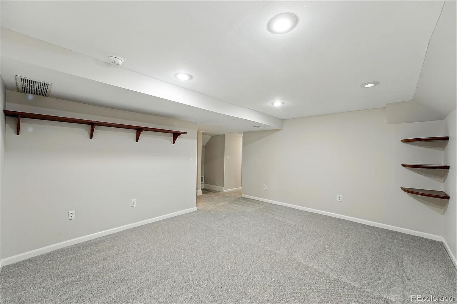The image showcases a finished basement with neutral gray carpeting and light gray walls. A long wooden shelf with supports is mounted on one wall, while three smaller shelves are on the opposite wall. Recessed lighting illuminates the space, creating a clean and versatile area.