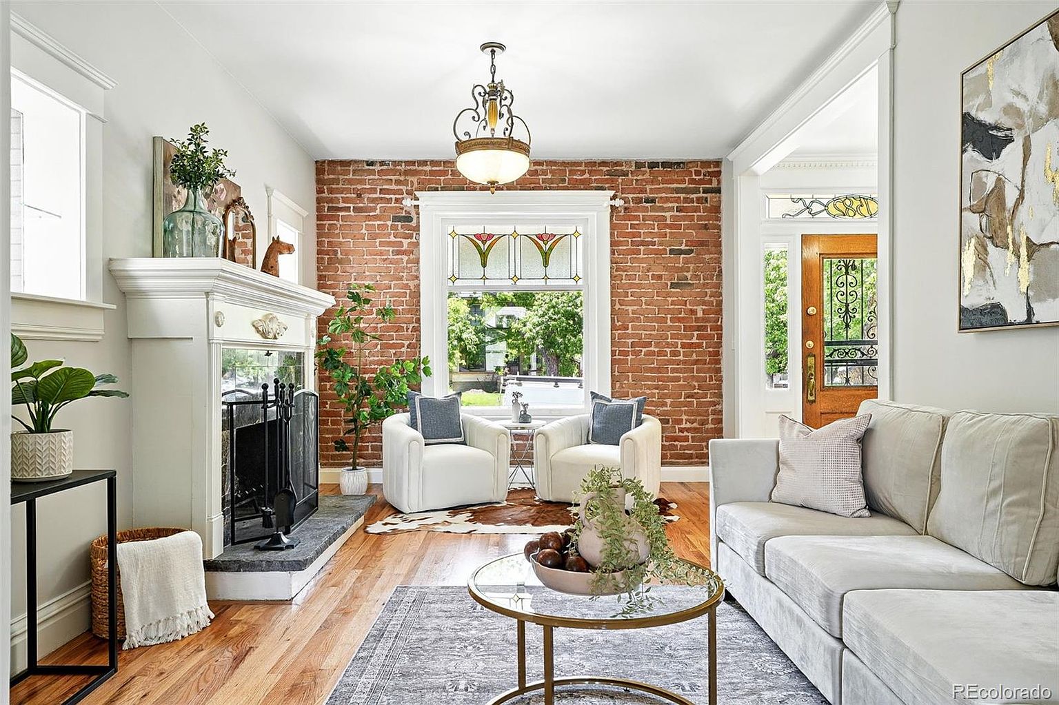 This is an inviting living room featuring a brick accent wall with a large window, two white armchairs, and a light gray sofa. A white fireplace mantel adds a classic touch, while a glass-topped coffee table sits atop a patterned rug. The room is well-lit and appears cozy and stylish, perfect for relaxation and entertaining.