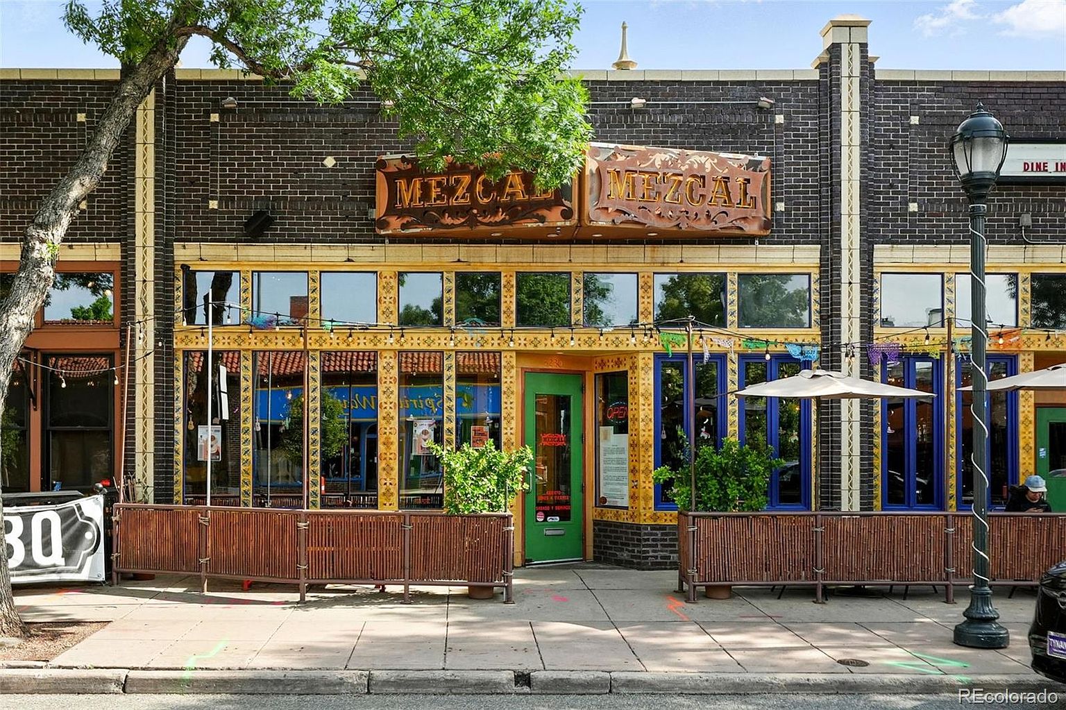 This is a front exterior view of a commercial building, likely a restaurant named "Mezcal", as indicated by the prominent signage. The building features a brick facade with decorative yellow trim around the windows and doors. An outdoor patio area is set up in front, enclosed by a wooden fence, suggesting a welcoming and vibrant atmosphere.