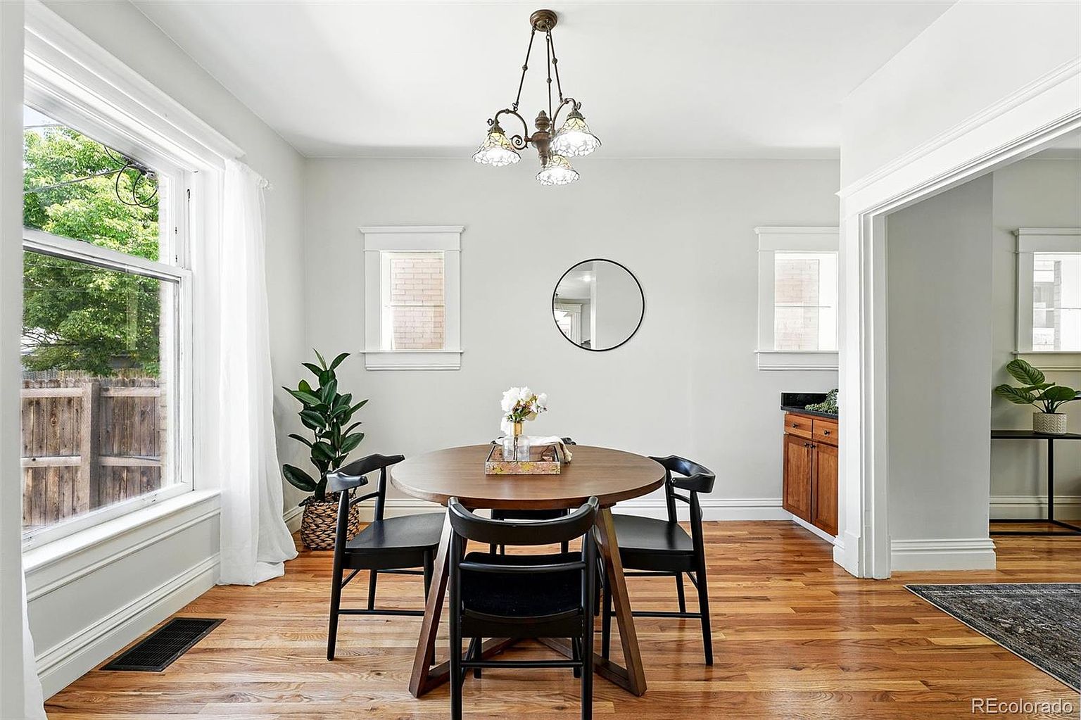 This is a dining room featuring a round wooden table with four black chairs, illuminated by a vintage-style chandelier. Natural light floods the room through a large window, complemented by two smaller windows on the adjacent wall. A round mirror hangs on the wall, adding depth to the space, and a plant adds a touch of greenery.