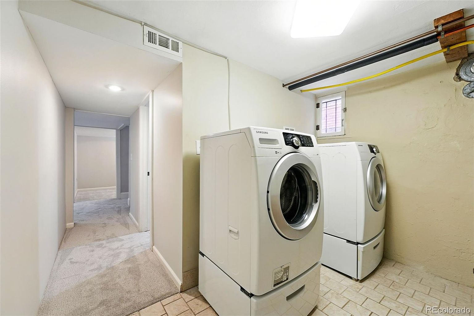 This is an interior shot of a laundry room featuring a Samsung front-load washer and dryer set, both in white, with a hallway visible in the background. The room has tiled flooring and beige walls, with a small window providing natural light. The overall impression is a functional and well-maintained laundry space.