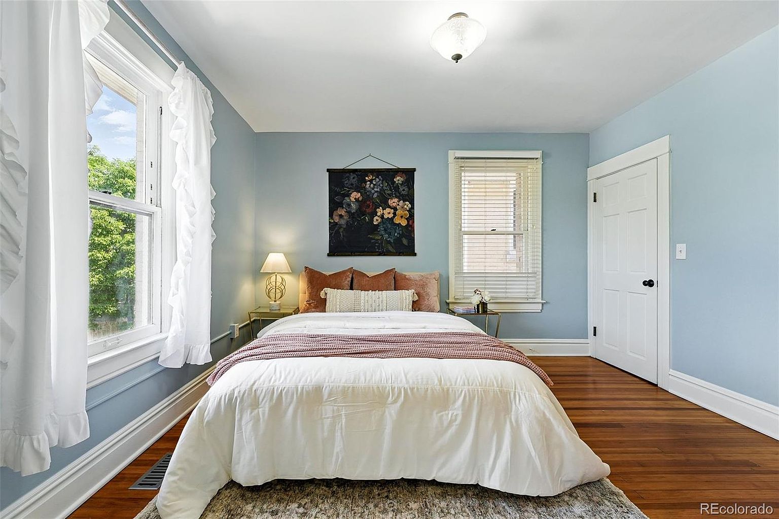 This is a cozy bedroom featuring a queen-sized bed with white bedding and a mauve throw. The walls are painted a calming blue, complemented by white trim and hardwood floors. A floral tapestry hangs above the bed, adding a touch of elegance, while natural light streams in through the window with white curtains.