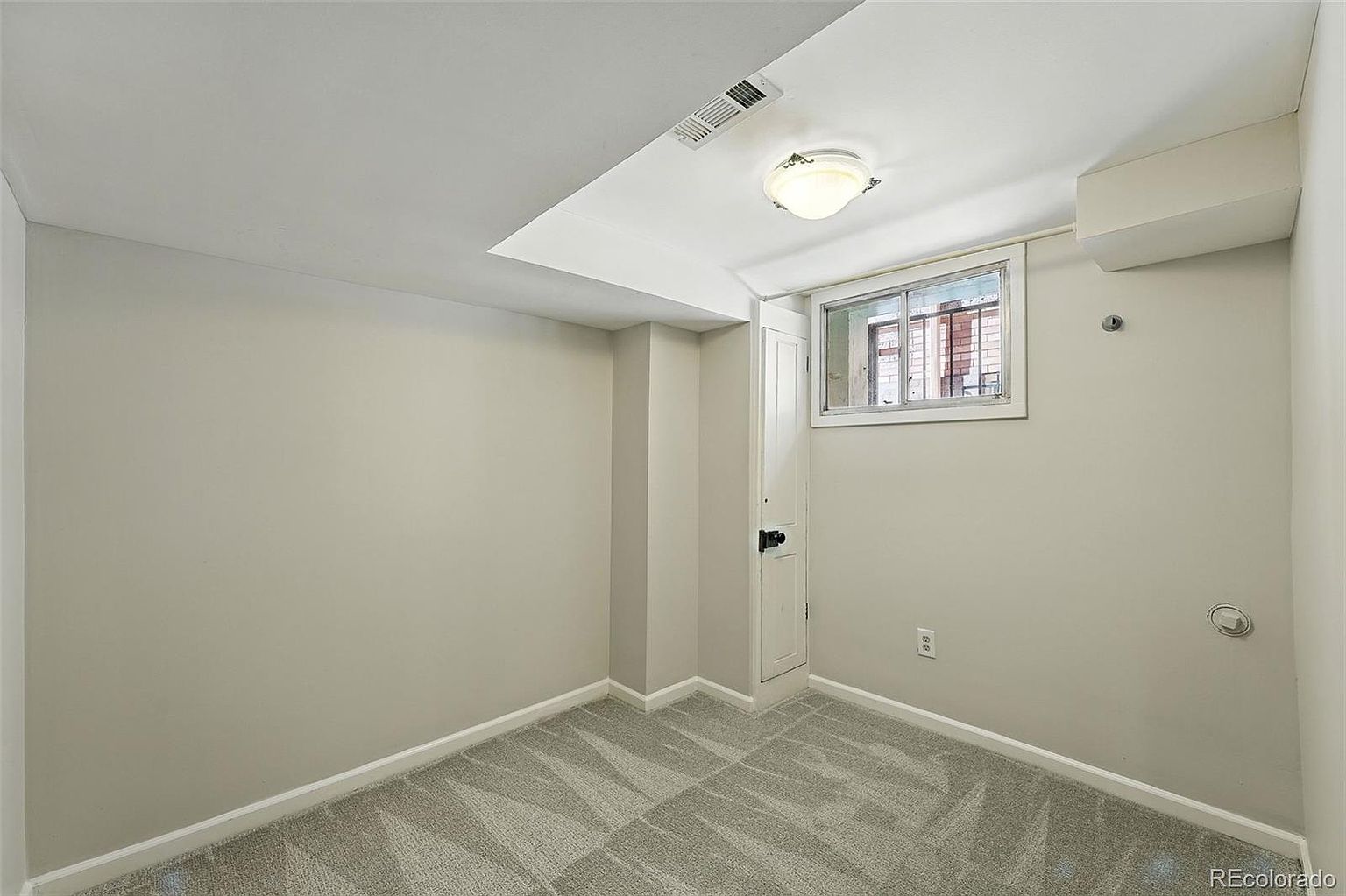 This is a simple, yet clean and functional bedroom. The room features neutral-colored walls, a gray patterned carpet, and a small window that provides natural light. A small closet is present, and the overall impression is one of a tidy and well-maintained space.