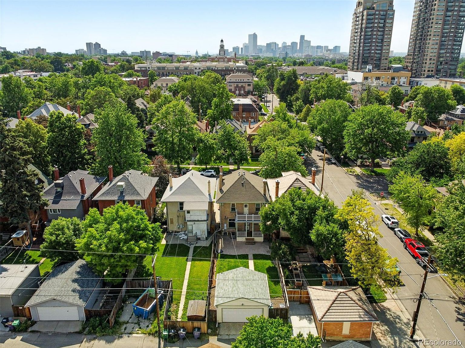 This aerial view showcases a neighborhood with well-maintained homes, lush green trees, and a cityscape in the background. The houses feature varied architectural styles, with neatly manicured lawns and detached garages. The perspective gives a sense of community and urban living.