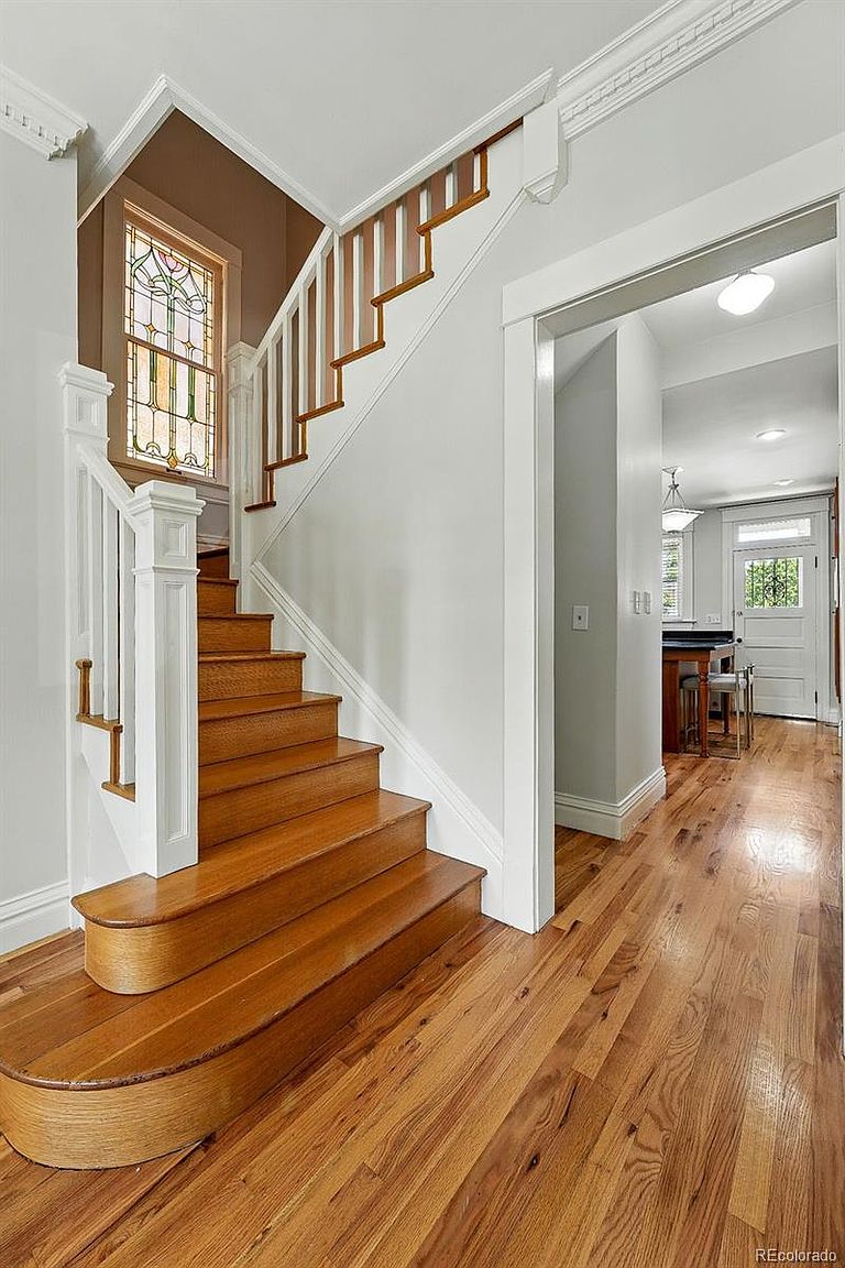 This interior shot showcases a well-lit hallway and staircase. The wooden stairs feature a curved bottom step and lead to an upper level, complemented by a white banister and a stained glass window. The hardwood flooring adds warmth, while the white walls and trim create a clean, classic aesthetic.