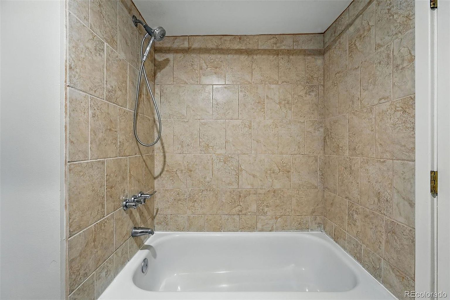 This is a bathroom featuring a white bathtub with tiled walls. The tiles are a neutral beige color with a subtle pattern. A shower head and faucet are visible, and the overall impression is clean and functional, suitable for a guest or secondary bathroom.