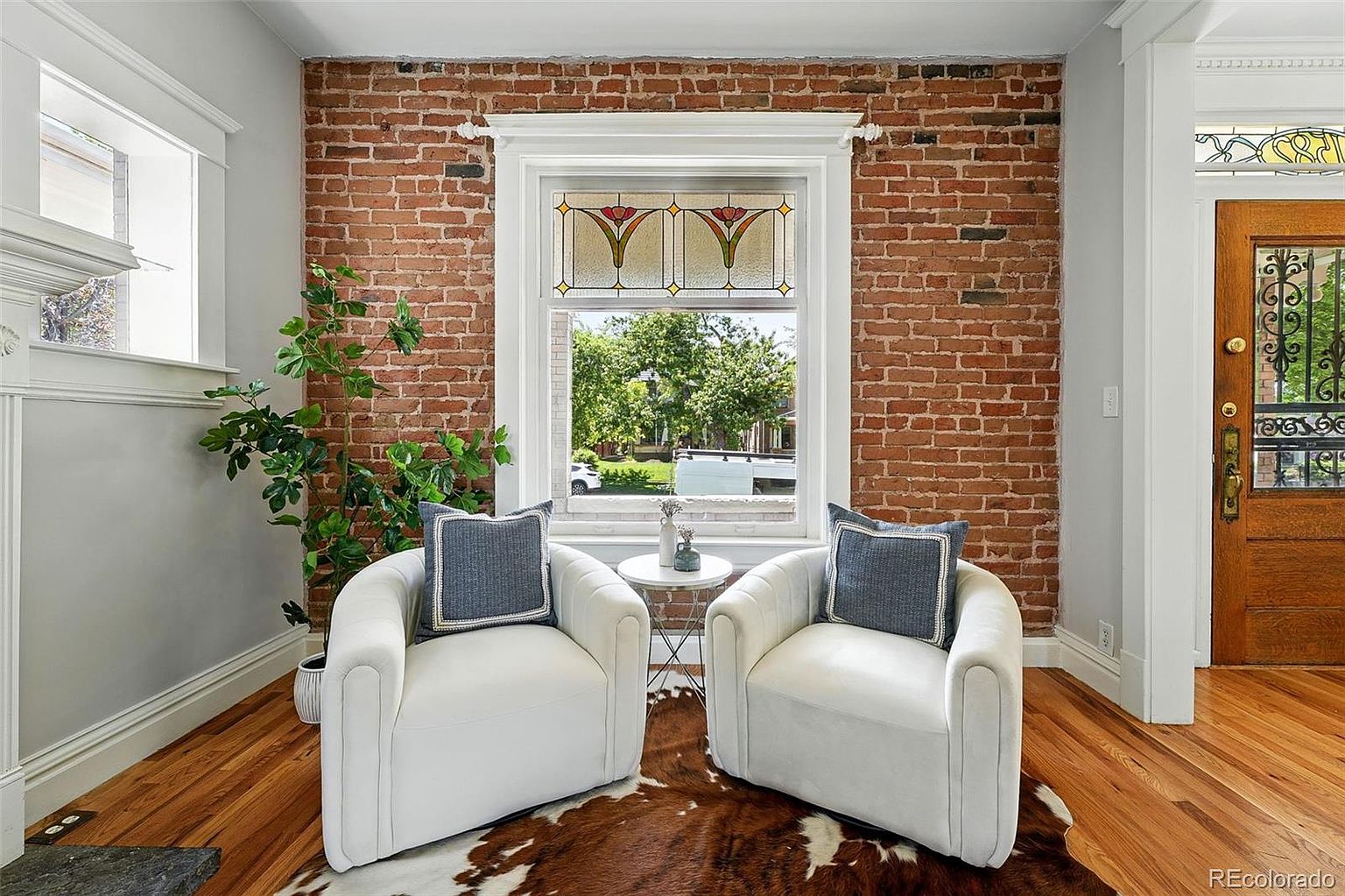 This interior shot showcases a cozy living room setup with two white armchairs flanking a small round table, all situated on a cowhide rug. A brick wall with a decorative window adds character, while a leafy plant brings a touch of nature to the space. The hardwood floors and natural light create a warm and inviting atmosphere.