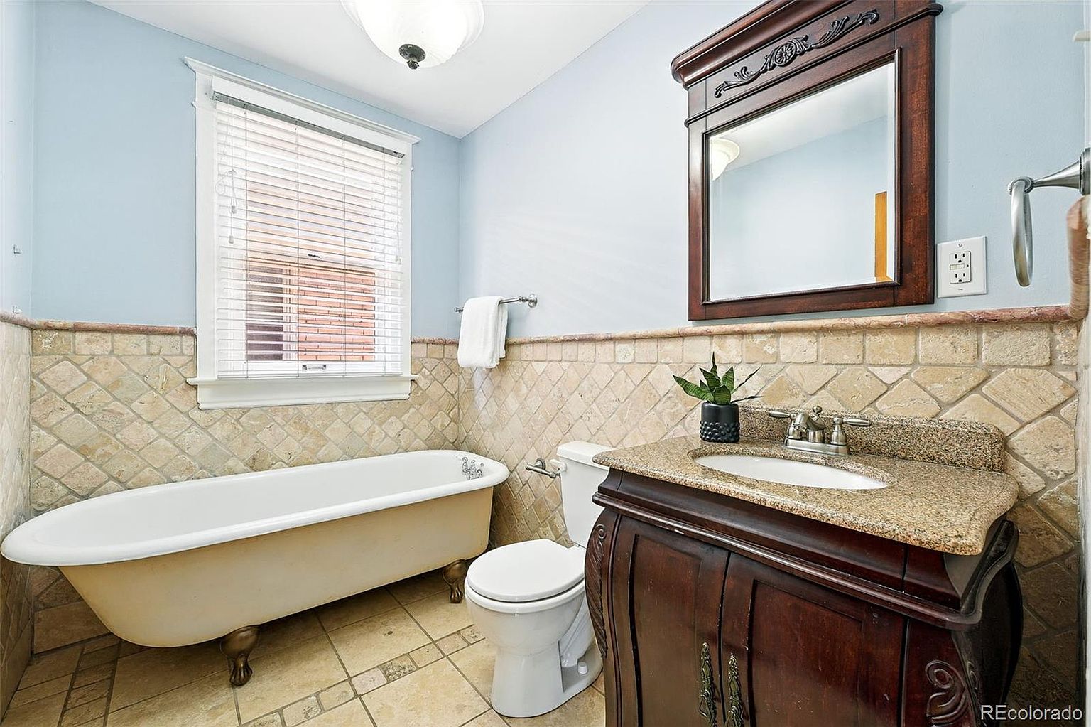 This is a bathroom featuring a clawfoot bathtub positioned beneath a window with blinds, a toilet, and a dark wood vanity with a granite countertop. The walls are light blue above a tiled wainscoting, and a framed mirror hangs above the vanity. The overall style is classic and well-maintained.