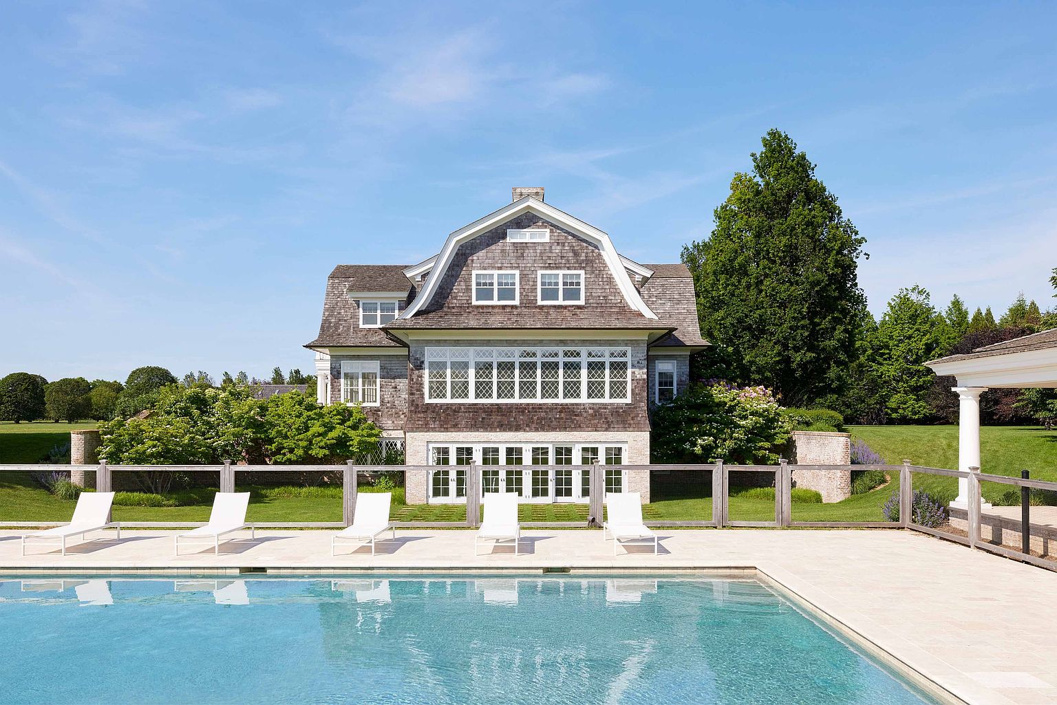This image showcases the rear exterior of a large, shingle-style home with a swimming pool in the foreground. The house features a gambrel roof, multiple windows, and a neutral color palette. White lounge chairs are arranged poolside, suggesting a relaxing outdoor living space, and the well-maintained lawn and landscaping enhance the property's appeal.