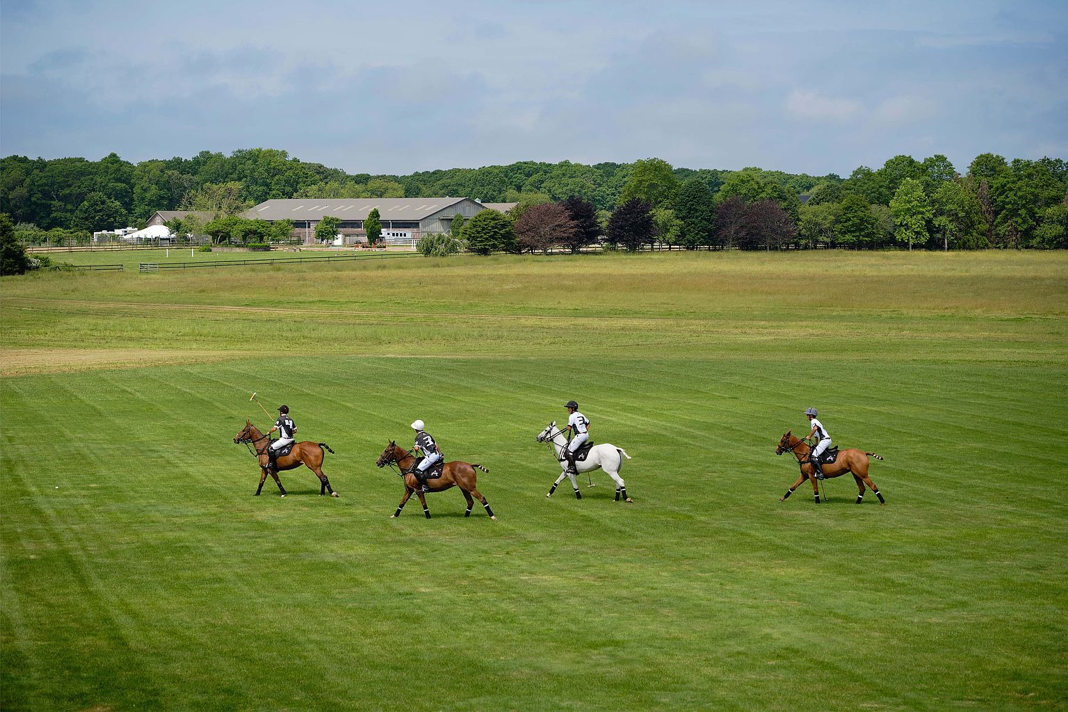 This expansive exterior shot showcases a meticulously maintained green lawn stretching towards a large building complex in the background, possibly stables or barns. Four polo players on horseback are actively engaged in a game, adding a dynamic element to the scene. The image conveys a sense of luxury, space, and recreational opportunities, ideal for a high-end real estate listing.