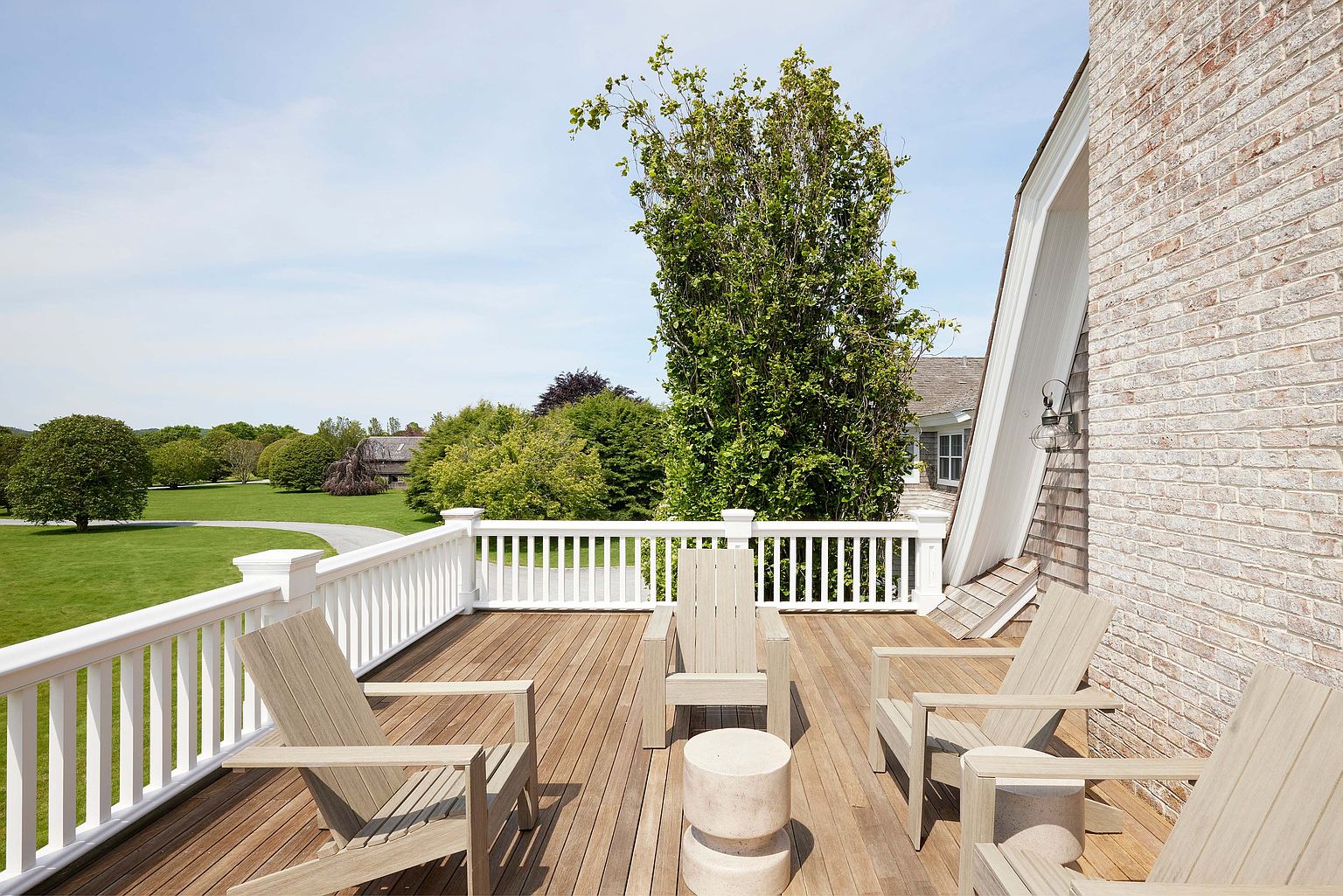 This image showcases a charming outdoor deck area, featuring wooden decking and a white railing that offers a view of a lush green lawn and mature trees. Several light-colored wooden Adirondack chairs are arranged around a small, cylindrical side table, creating an inviting space for relaxation and outdoor enjoyment. The deck is adjacent to a brick building, adding architectural interest to the scene.