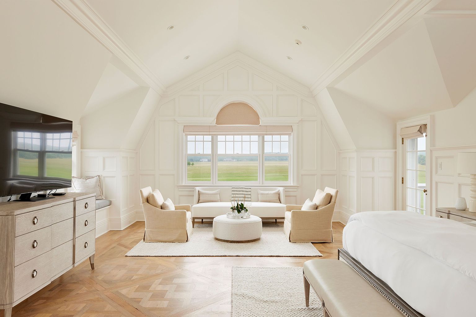 This is a bright and airy primary bedroom featuring a vaulted ceiling with detailed paneling and trim. The room is furnished with neutral-toned seating, including armchairs and a settee, arranged around a round ottoman on a light-colored rug. A large window provides ample natural light and a view of the landscape, while a TV and dresser are positioned on the left side of the room.