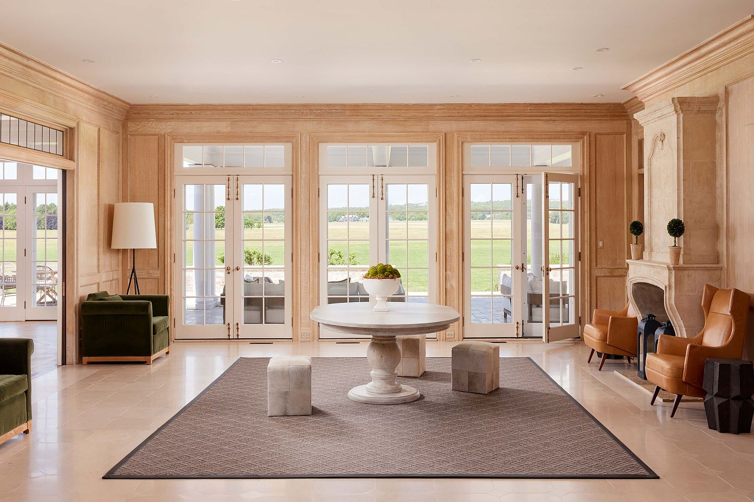 This is a spacious and elegant living room featuring a neutral color palette with light wood paneling and flooring. The room is well-lit with natural light streaming through multiple French doors that lead to an outdoor area. A large round table with decorative elements sits in the center of the room, complemented by comfortable seating arrangements including armchairs and ottomans, creating a luxurious and inviting atmosphere.