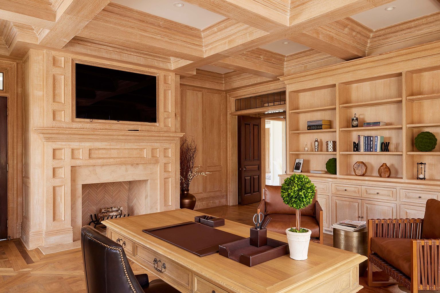 This is an elegant home office featuring wood paneling, a coffered ceiling, and built-in bookshelves. A large wooden desk with leather accents sits in the center of the room, complemented by a leather chair. A fireplace with a mounted television above it adds a focal point, creating a sophisticated and comfortable workspace.