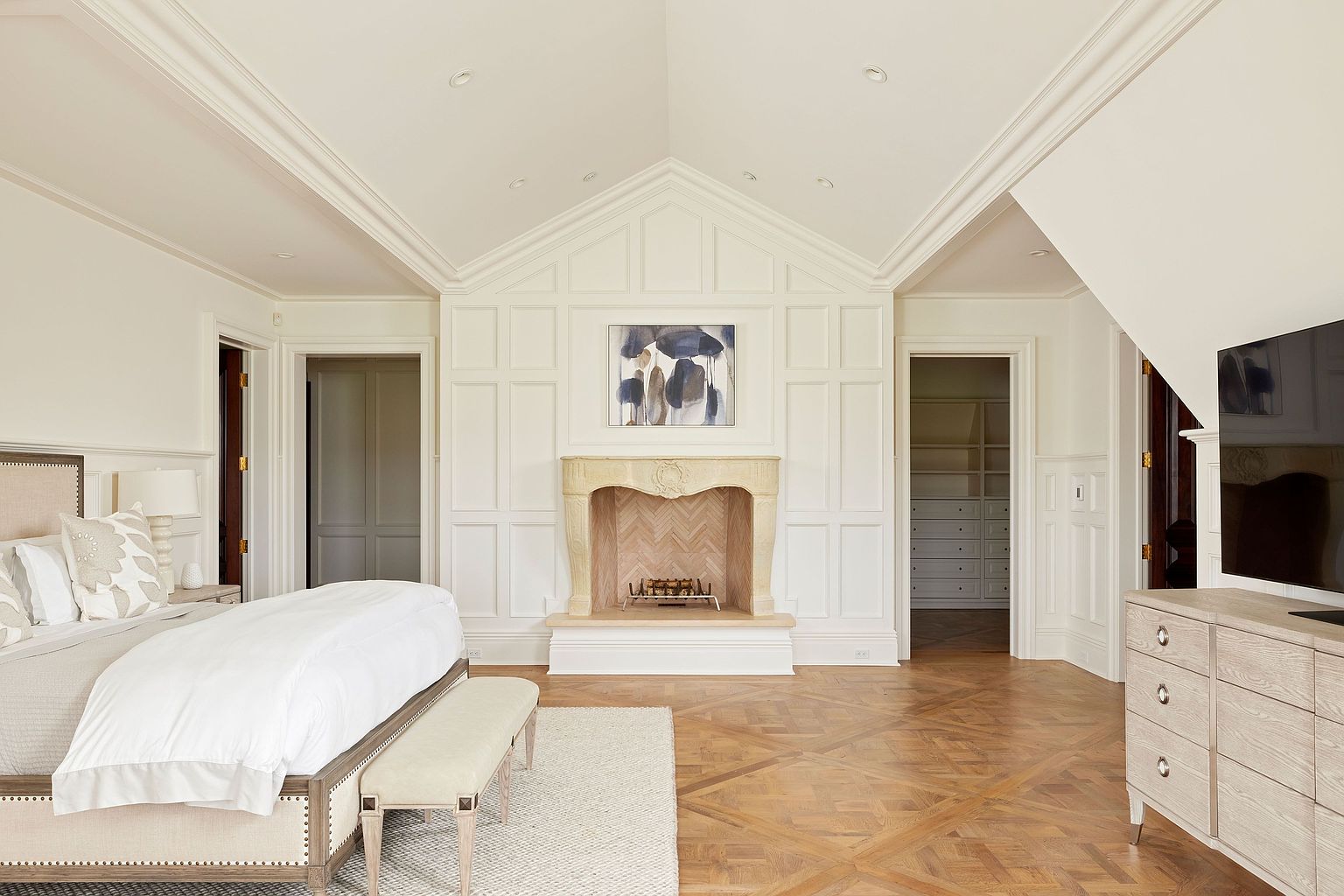 This is a primary bedroom featuring a neutral color palette and a sophisticated design. The room includes a large bed with a padded headboard, a fireplace with a decorative mantel, and hardwood floors laid in a herringbone pattern. There are doorways leading to other rooms, and a TV is mounted on a dresser. The perspective is a wide shot, showcasing the room's layout and architectural details.