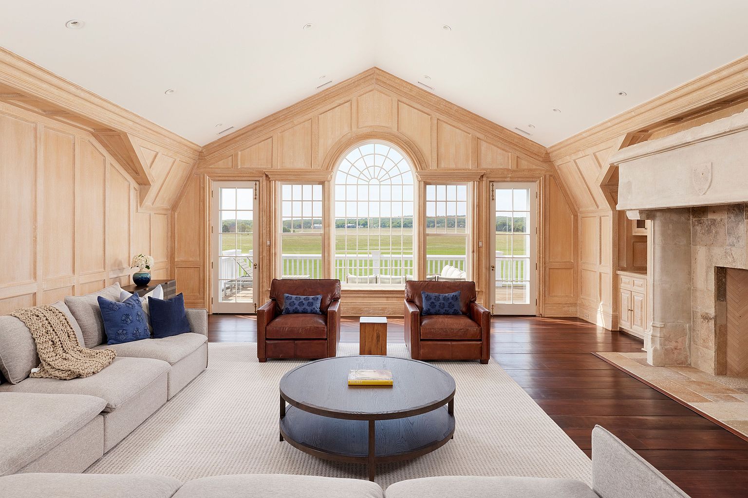 This is a spacious living room featuring light wood paneling, a high vaulted ceiling, and a large arched window offering a view of a green field. The room is furnished with a gray sectional sofa, two leather armchairs, and a round coffee table. The dark wood flooring contrasts with a light-colored area rug, and a stone fireplace adds a touch of rustic elegance.