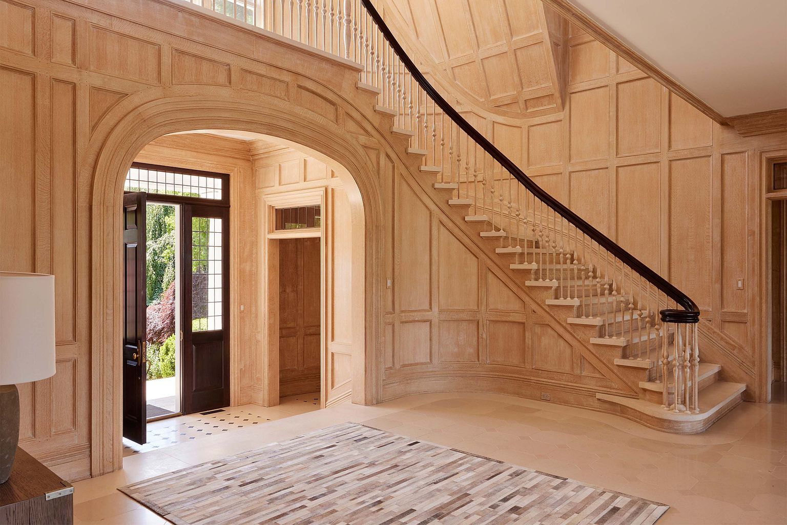 This grand foyer showcases a stunning staircase with a dark wood handrail and light wood spindles, complemented by paneled walls and arched doorways. The flooring is a light-colored tile, and a patterned rug adds warmth to the space. The overall impression is one of elegance and timeless design.