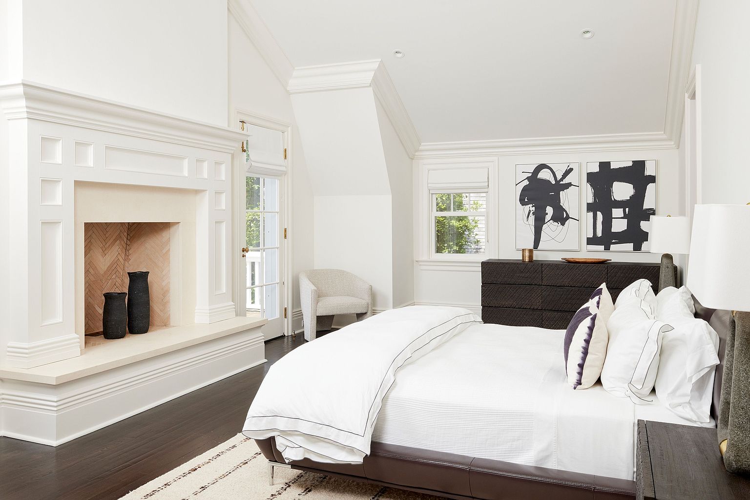 This is a bright and airy primary bedroom featuring a fireplace with decorative vases, a dark wood dresser with abstract art above, and a bed with white linens and a brown leather frame. The room has a sophisticated and calming atmosphere, enhanced by the natural light from the window and the neutral color palette. The dark wood floors add a touch of elegance.