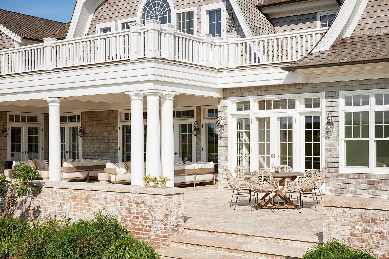 This image showcases a luxurious outdoor patio area, featuring a dining set with woven chairs and a wooden table, complemented by comfortable lounge seating under a covered porch supported by elegant white columns. The patio is constructed with stone pavers and is partially enclosed by a brick wall, adding to the sense of privacy and sophistication. The overall impression is one of refined outdoor living and relaxation.