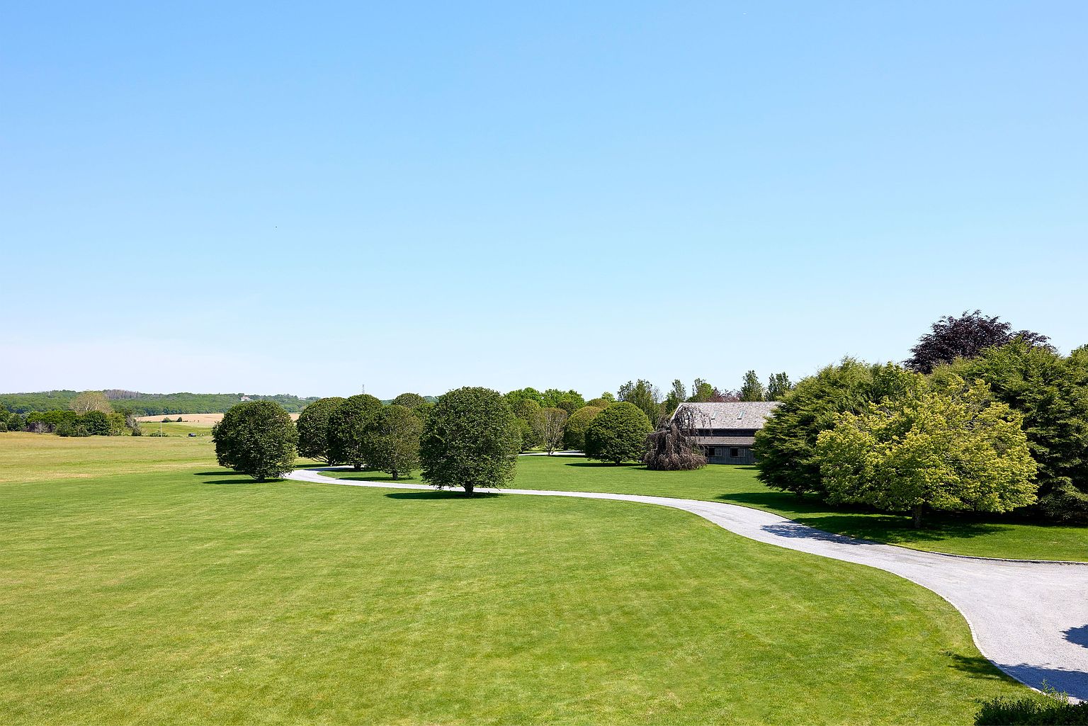 This aerial view showcases a sprawling green lawn leading to a charming house nestled among mature trees. A winding driveway adds to the property's appeal, suggesting privacy and a grand entrance. The clear blue sky enhances the sense of openness and tranquility, making it an ideal estate.