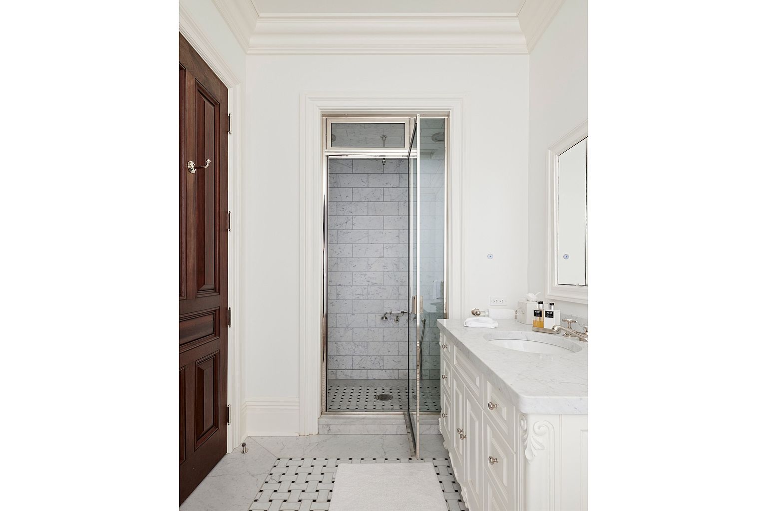 This is a bright and elegant primary bathroom featuring a marble-topped vanity with white cabinetry and classic hardware. A glass-enclosed shower with marble tile and a mosaic floor adds a luxurious touch. The flooring is a black and white mosaic pattern, and a dark wood door provides a contrast to the light color palette.