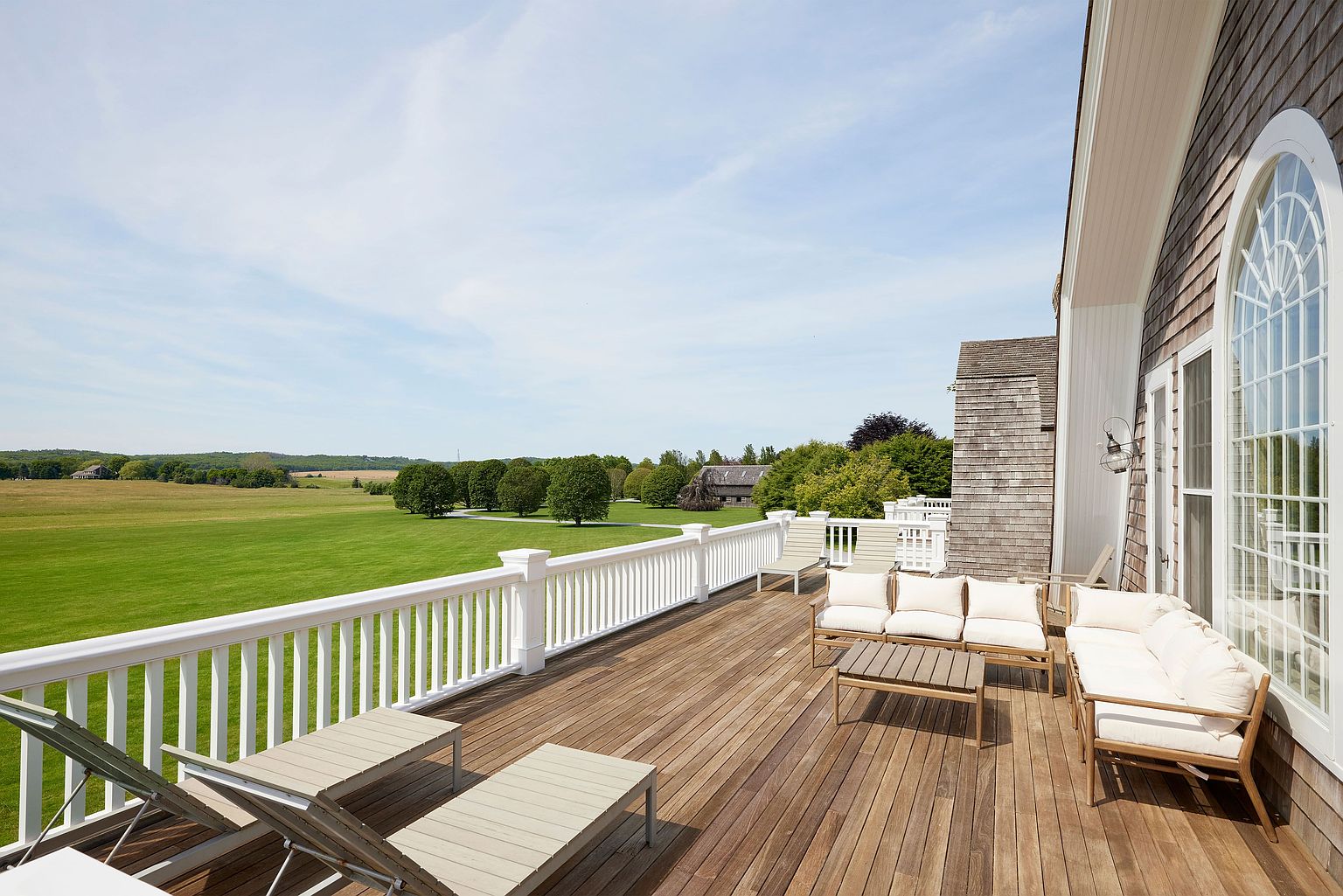 This image showcases a spacious wooden deck or balcony with comfortable outdoor furniture, including lounge chairs and a sofa set with white cushions. The deck overlooks a vast green lawn and distant trees, offering a serene and picturesque view. The architectural details of the house, such as the white railing and shingle siding, add to the property's charm.