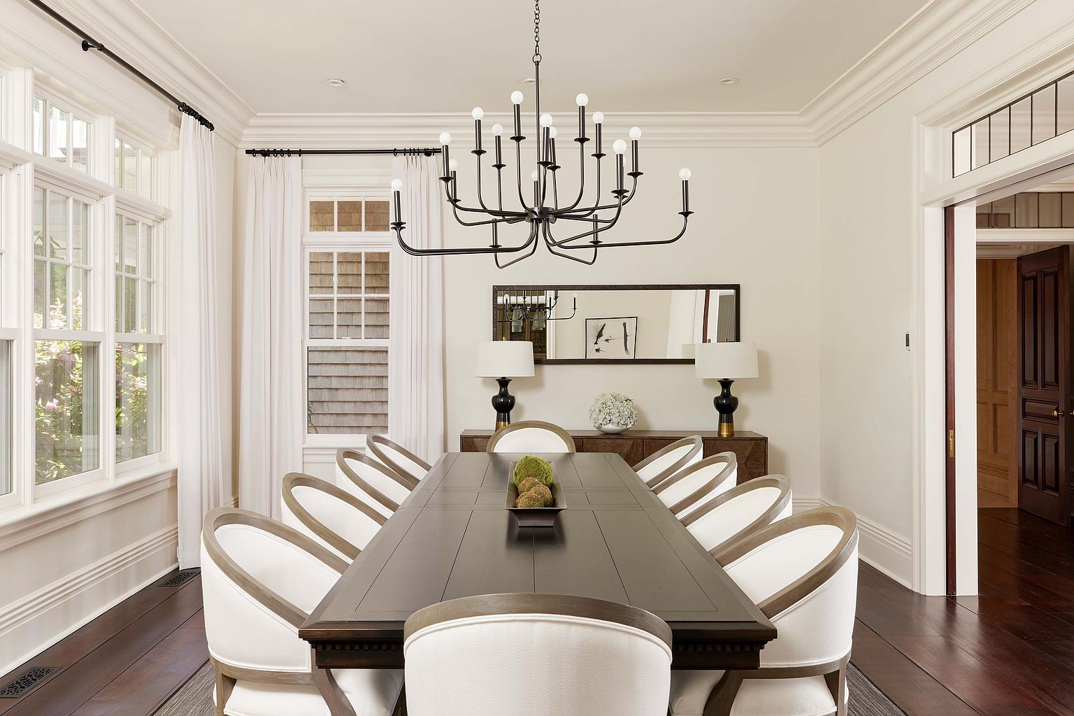 This is an interior shot of a dining room featuring a long, dark wood table surrounded by upholstered chairs with light wood frames. A modern chandelier hangs above the table, and a sideboard with lamps and a mirror is positioned against the wall. The room is well-lit with natural light from the windows, creating an elegant and inviting atmosphere.