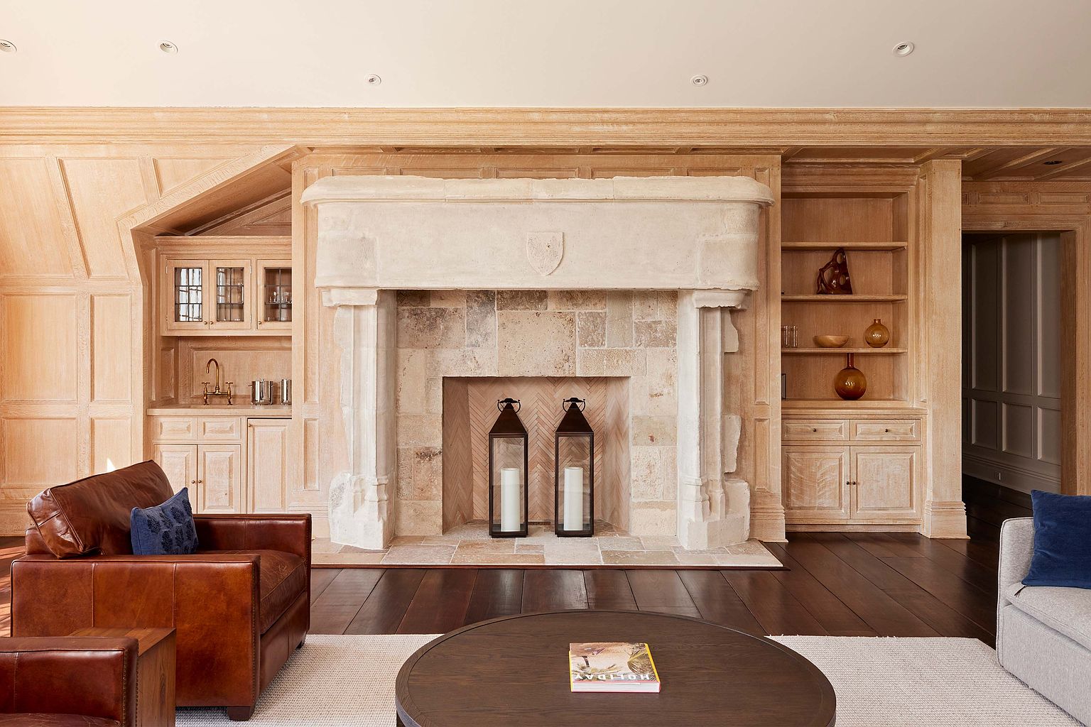 This living room showcases a grand stone fireplace flanked by custom built-in cabinetry, creating a focal point in the room. The dark hardwood floors contrast with the light-colored walls and furniture, adding depth and warmth to the space. A leather armchair and a round coffee table contribute to the room's inviting and sophisticated atmosphere.