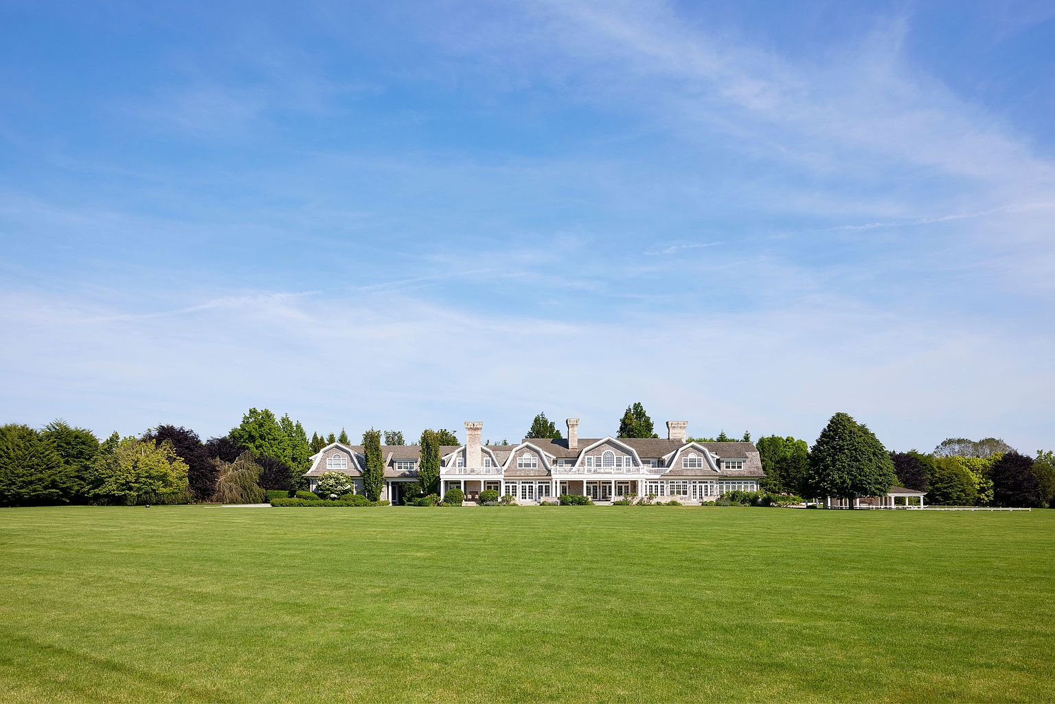 This image showcases the grand front view of a large, luxurious house with a sprawling green lawn. The house features multiple gabled roofs, chimneys, and a symmetrical facade with numerous windows and a covered porch. The well-manicured lawn extends to the front of the property, creating an impressive and inviting approach.