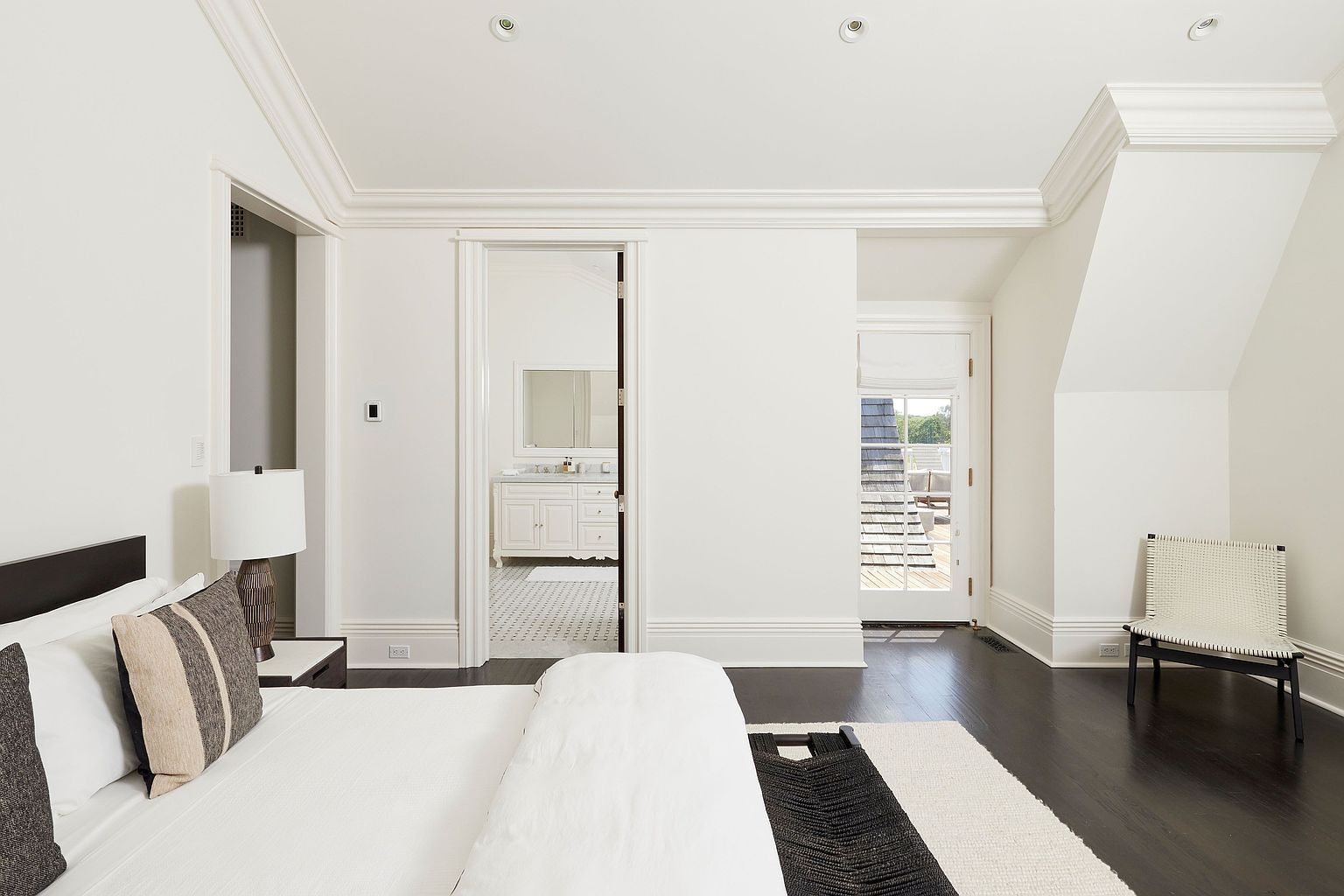 This is a bright and airy primary bedroom featuring white walls, dark hardwood floors, and elegant crown molding. The room includes a bed with white linens and decorative pillows, a doorway leading to a bathroom, and a door to a balcony. A woven chair and a black and white rug add texture and visual interest to the space.