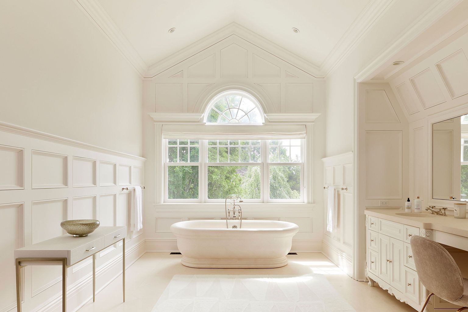 This is a luxurious primary bathroom featuring a freestanding bathtub positioned in front of a large window offering natural light and a view of greenery. The bathroom is decorated in a classic style with white paneled walls, a vanity with a sink, and a neutral color palette, creating a serene and elegant atmosphere. A console table with a decorative bowl adds a touch of sophistication.