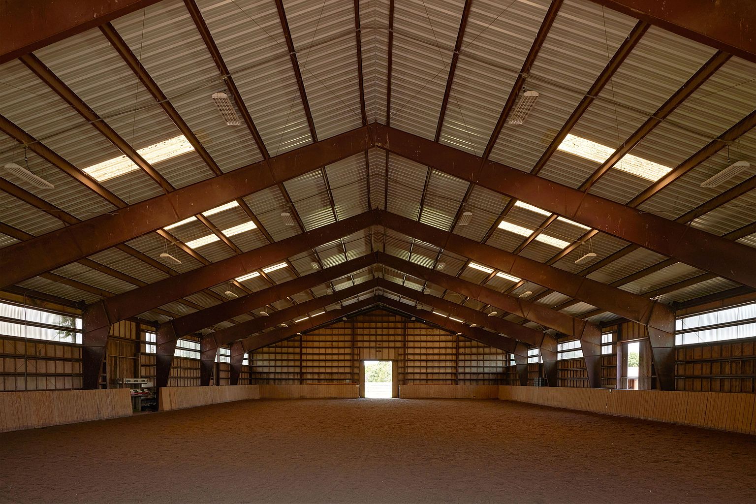 This is an interior shot of a large indoor riding arena. The arena features a high, peaked metal roof supported by steel beams, with skylights providing natural illumination. The riding surface is composed of sand or dirt, and the perimeter is lined with a low wooden wall.