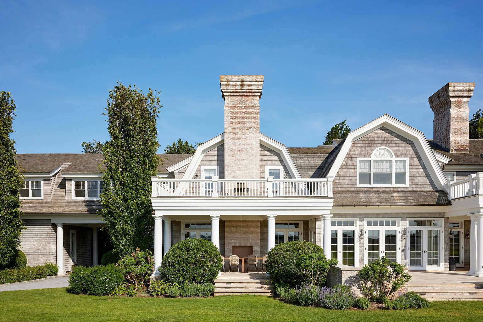 This is a rear view of a large, luxurious home with a shingle-style exterior. The house features a prominent brick chimney, a covered balcony with white columns, and multiple sets of French doors leading to a patio area. The well-manicured lawn and landscaping add to the property's appeal.