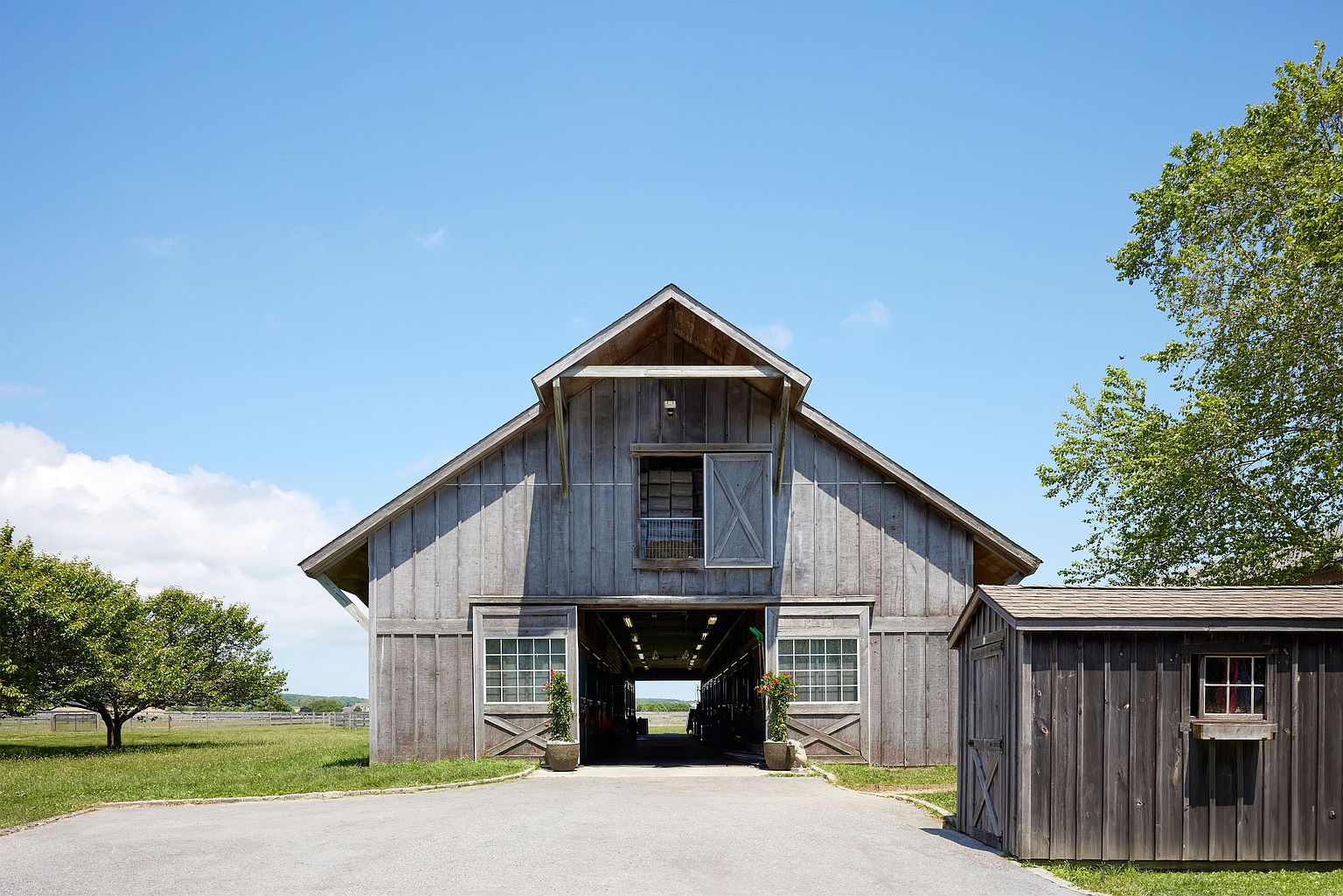 The image showcases the front view of a large, weathered gray barn with a prominent gable roof and wide sliding doors. Two windows flank the entrance, each adorned with potted plants. A smaller, matching shed is situated to the right, and a paved driveway leads up to the barn, set against a backdrop of a clear blue sky and green fields.