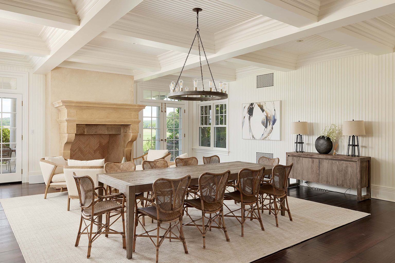 This is an elegant dining room featuring a long wooden table surrounded by wicker chairs, set on a large neutral rug. A stone fireplace adds a touch of warmth, while a modern chandelier hangs above the table. The room is well-lit with natural light and features a sideboard with lamps and artwork, creating a sophisticated and inviting atmosphere.
