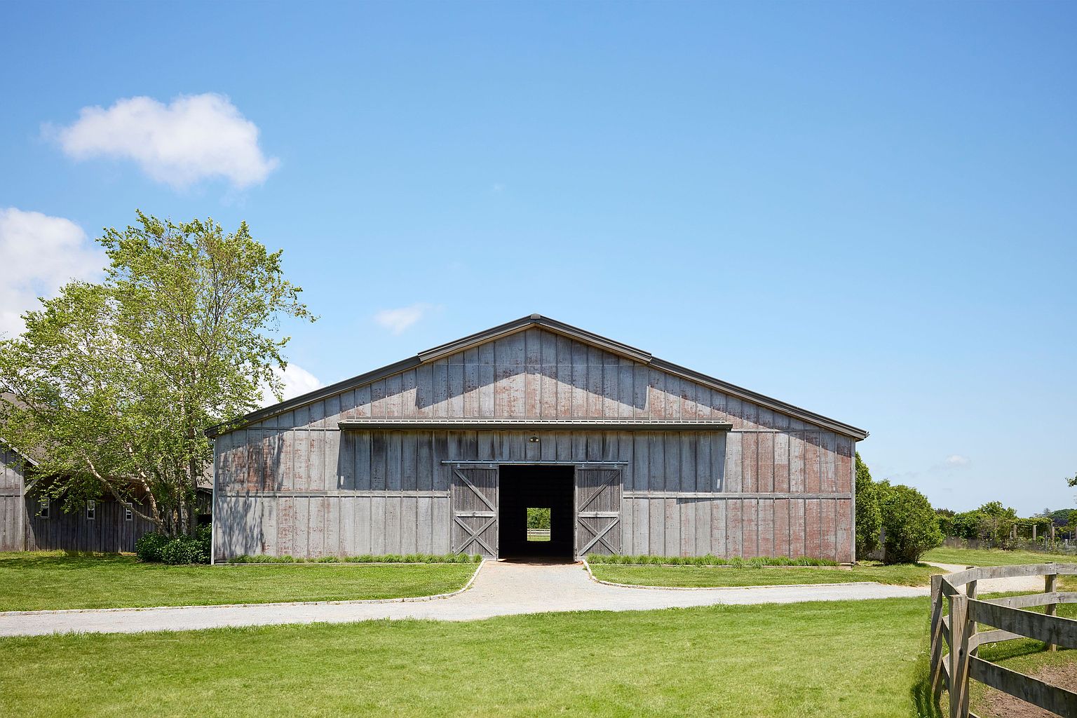 The image showcases the front view of a large, rustic barn-style building with weathered gray siding and a prominent gable roof. A gravel path leads to the barn's entrance, flanked by well-maintained green lawns. The structure exudes a sense of rural charm and spaciousness, making it an ideal property for those seeking a unique and character-filled home or event space.