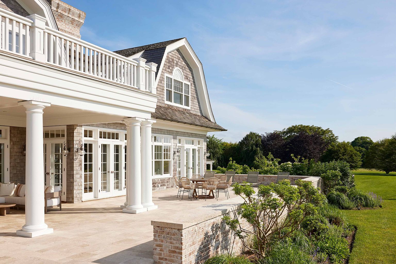 This image showcases a luxurious patio or deck area of a high-end home. The space features a stone-tiled floor, elegant white columns supporting an upper balcony, and a dining set for outdoor entertaining. A low brick wall borders the patio, with lush landscaping and mature trees in the background, creating a serene and private outdoor living space.